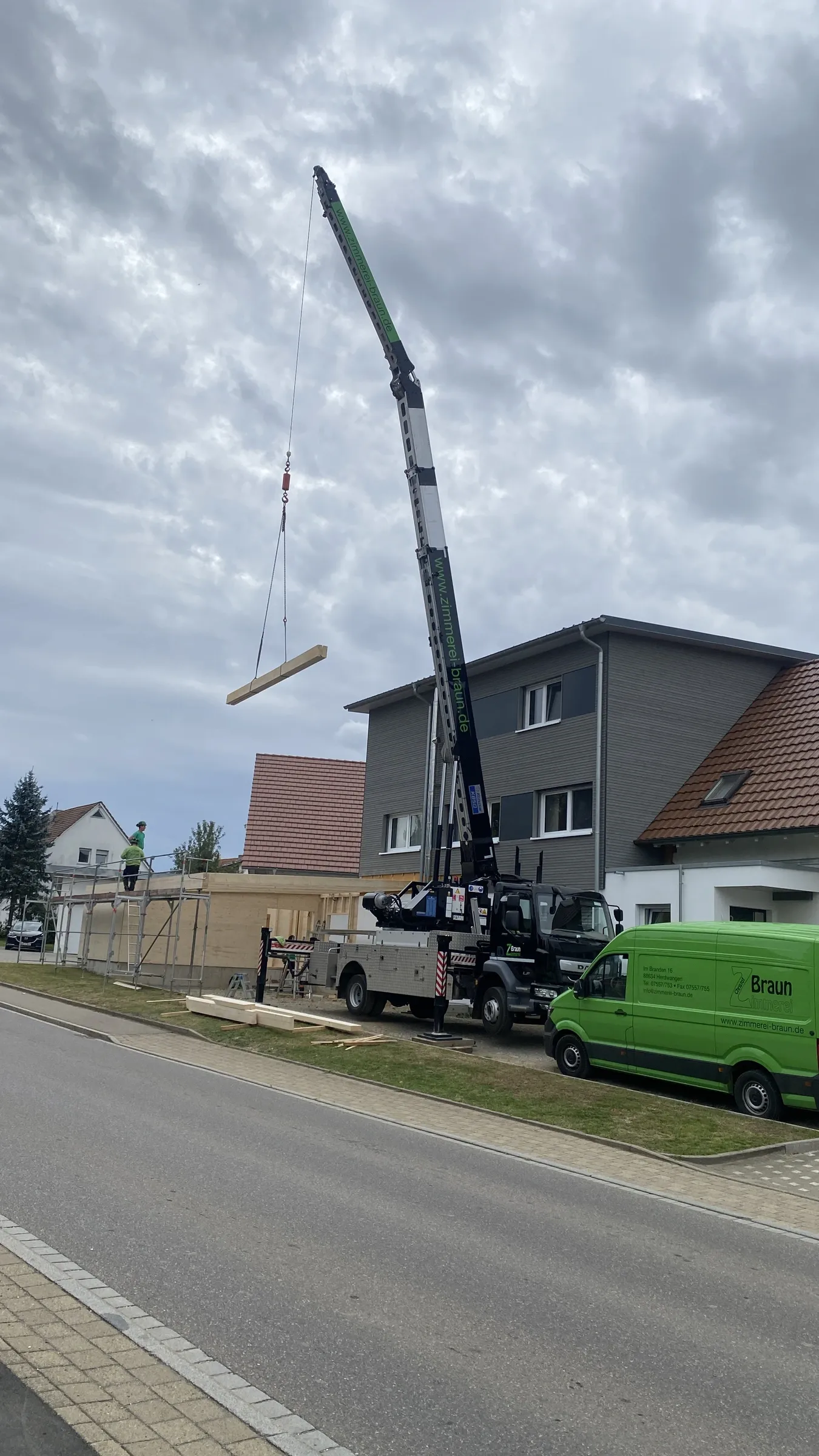 Abgestützter Autokran am Straßenrand schwenkt einen Holzbalken zum eingerüsteten Rohbau