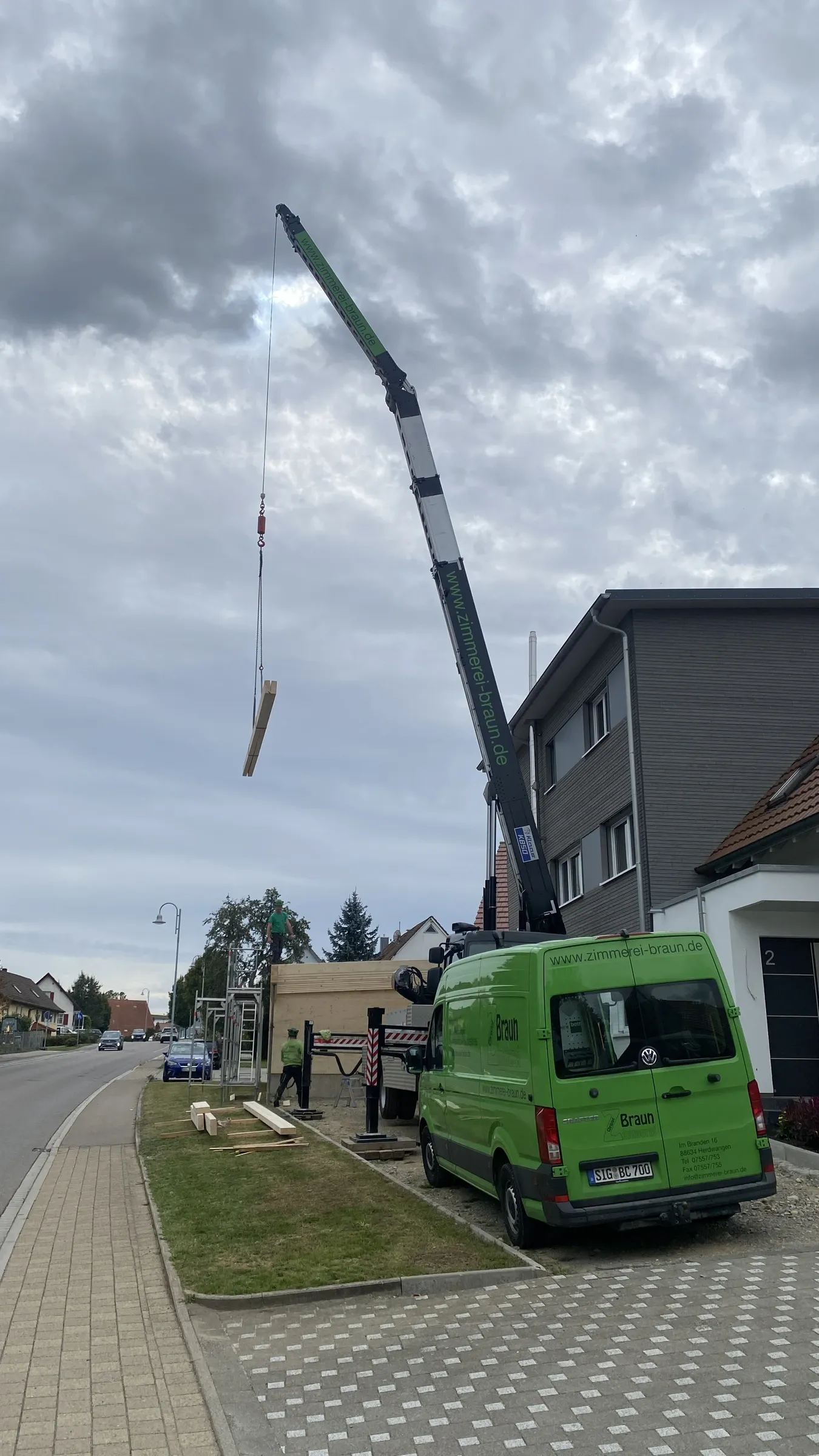 Autokran hebt einen Holzbalken über eine Baustelle, davor der grüne Transporter der Zimmerei Braun